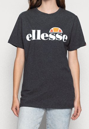 Persoon draagt donkergrijs T-shirt met witte "ellesse" tekst en oranje halvemaanvormig logo, gecombineerd met lichtblauwe spijkerbroek.
