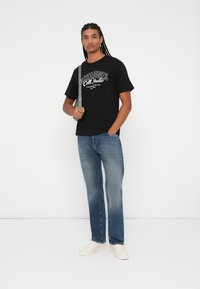 T-shirt preta de gola redonda com texto gráfico branco, combinada com jeans de lavagem azul clara de corte solto e sapatos de slip-on na cor creme.