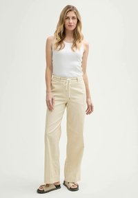 Pantalons larges beige clair avec une taille à cordon, fabriqués dans un tissu doux, associés à un débardeur blanc simple et des sandales beiges.