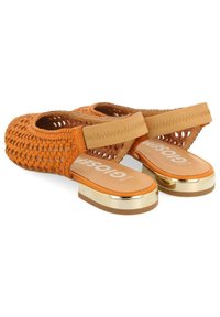 Sandalias de color naranja tejidas con una correa beige en el tobillo, suela plana y un acento dorado en el tacón, con un diseño de espalda abierta y forma redondeada.