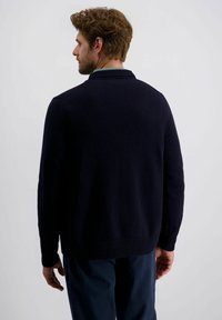 Mann mit kurzem lockigem Haar und Bart, der einen dunkelblauen Pullover und eine blaue Hose trägt, von hinten vor einem schlichten Hintergrund gesehen.