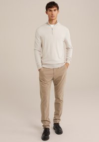 Svetlo bež zip-up pulover z dolgimi rokavi in ribanimi manšetami, v kombinaciji z bež hlačami in črnimi čipkastimi čevlji. Preprost, sodoben dizajn.