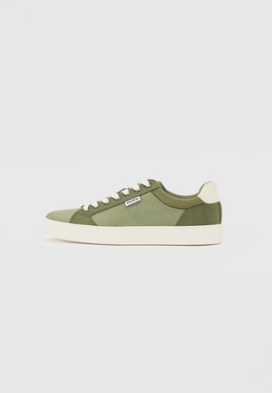Grønne lerrets sneakers med kremfargede detaljer, avrundet tå og hvit gummisåle; har sømdetaljer og en etikett på siden.