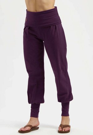Femme portant un pantalon violet large taille haute avec des chevilles ajustées et des sandales marron, debout devant un fond blanc uni.