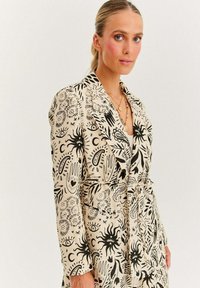 Béžový blazer s černými abstraktními vzory, tvarovaný design, klopové límce a zavázaný pás. Texturovaná látka s uměleckými motivy.