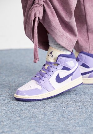 Personne portant des chaussettes blanches et des baskets Air Jordan violet-blanc, avec un pantalon mauve noué à la cheville, debout sur un tapis bleu.