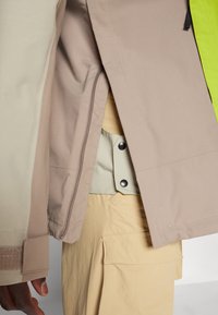 Beige og lysegrønn jakke med kontrasterende neongrønn aksent, frontglidelås og trykknapper. Teksturert stoff med synlige sømmer.