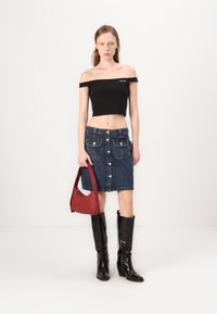 Top cropped nero off-the-shoulder, gonna di jeans con due tasche frontali, borsa rossa e stivali al ginocchio neri lucidi.
