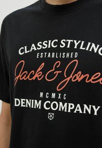 Melnā kokvilnas T-kreklā ir drosmīgs balts un oranžs teksts: "Klasiska stilistika", "Jack & Jones", "Izveidots" un "Denim Company". Klasisks apaļais kakls.