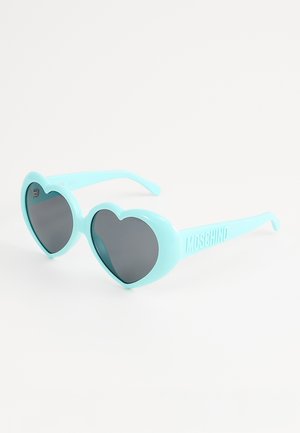 Occhiali da sole blu a forma di cuore con lenti scure e marchio "Moschino" sulle aste, posati su una superficie bianca.