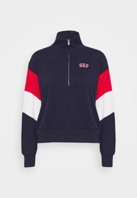Felpa navy con colletto alto, zip a metà, maniche a righe diagonali rosse e bianche, e piccolo logo "GAP" rosso sul petto. Tessuto misto cotone.