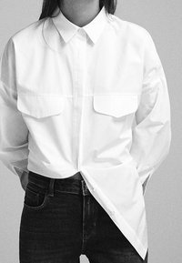 Camicia bianca con colletto e due tasche frontali, realizzata in un tessuto liscio; abbinata a jeans in denim scuro, creando un look a contrasto.