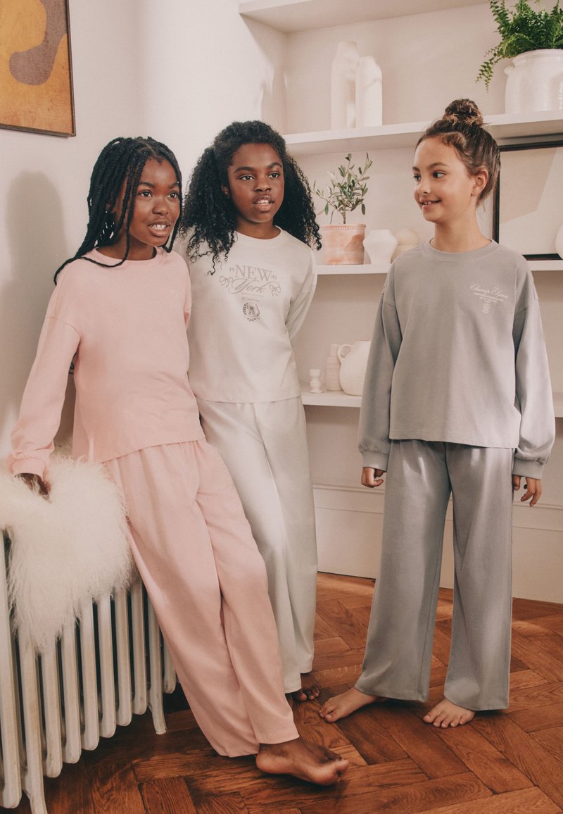 Drei Mädchen in pastellfarbener Loungewear stehen barfuß auf einem Holzboden in einem gemütlichen Raum mit Regalen und Pflanzen im Hintergrund.