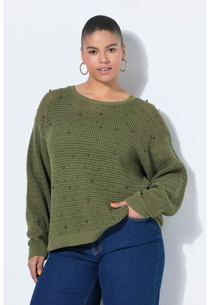 Mladá žena s krátkými vlasy nosí olivově zelený pletený svetr s korálkovými detaily a modré džíny, stojící před bílou zdí.