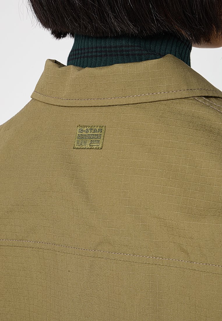 Olivgrüne strukturierte Jacke mit einem quadratischen Muster, ausgestattet mit einem G-Star-Label am Kragen und einem dunkelgrünen gerippten Rollkragenpullover darunter.