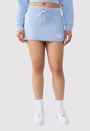 Femme portant une mini-jupe à cordon bleu clair avec un ourlet effiloché, un logo blanc, des chaussettes blanches et des baskets blanches, debout devant un fond uni.