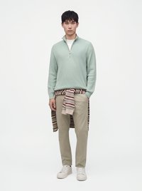 Jeune homme portant un pull vert clair à fermeture éclair quart, un pantalon beige, des baskets blanches, avec un pull rayé noué autour de la taille, debout devant un fond blanc.