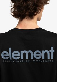 Camiseta de algodón negra con gran texto en gris claro que dice "element SKATEBOARD CO. WORLDWIDE" en la parte trasera. Cuello redondo.
