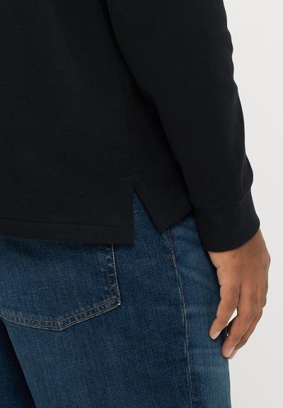 Μαύρο μακρυμάνικο πουκάμισο με ριμπ υφή, με πλάγιες σχισμές στο τελείωμα. Συνδυασμένο με σκούρο τζιν παντελόνι, που δείχνει πίσω τσέπη.