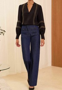 Blusa negra con mangas abullonadas y tachuelas metálicas, combinada con pantalones vaqueros oscuros de pierna ancha y cintura alta. Tela suave y diseño a medida.