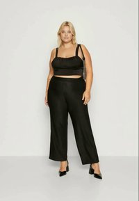 Top preto cortado com detalhes franzidos, combinado com calças largas pretas. A modelo usa saltos altos pretos com bico e segura uma pequena mala preta.