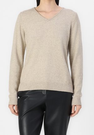 Femme portant un pull en maille beige clair à col en V et un pantalon en cuir noir, debout devant un fond blanc uni.
