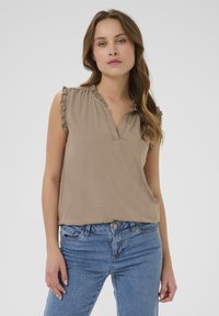 Un haut sans manches beige avec un col froncé et un plastron de boutons, fabriqué en tissu doux, associé à un jean bleu en denim.