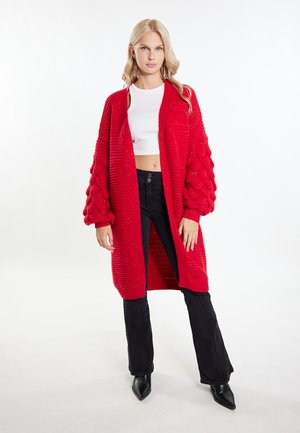 Roter gestrickter Cardigan mit Rippenstruktur und Puffärmeln, getragen über einem weißen Cropped-Top und kombiniert mit schwarzen Schlaghosen und spitzen Absätzen.