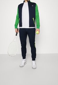 Träningskläder som består av en marinblå jacka med gröna ärmar, en vit skjorta, marinblå joggers med gröna detaljer och vita sneakers. Håller en tennisracket och en boll.