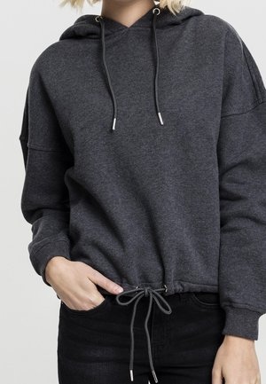 Personne portant un sweat à capuche gris foncé avec des cordons et un pantalon noir, avec une main dans la poche et l'ourlet du sweat noué en nœud.
