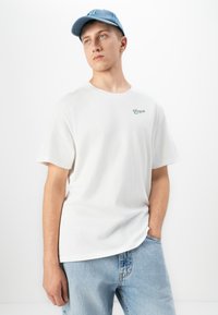 Young man in light blue cap, white Pompei logo T-shirt, and light blue jeans, standing with one hand in pocket against white background.