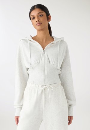 Donna con capelli scuri raccolti in una coda di cavallo che indossa una felpa con cappuccio grigio chiaro con zip e pantaloni della tuta coordinati con coulisse su uno sfondo semplice.