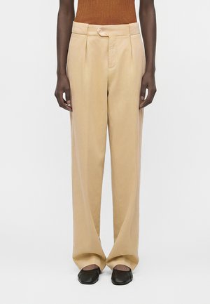 Άτομο που φορά μπεζ παντελόνια με φαρδιά μπατζάκια, καφέ ριμπ πουκάμισο και μαύρα παπούτσια με τετράγωνη μύτη, στέκεται μπροστά σε απλό φόντο.