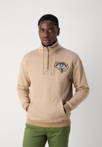 Beige zip-up sweatshirt with high collar and embroidered design on chest. Features a front pocket and ribbed cuffs and hem.