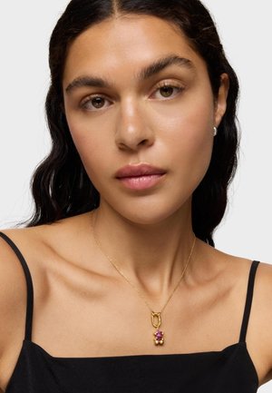 Mujer de cabello oscuro que lleva una blusa negra de tirantes finos y un collar dorado con un colgante de piedra preciosa púrpura, sobre un fondo claro y liso.