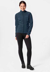 Navy softshell jacket with full zipper, high collar, and long sleeves, paired with black fitted leggings and black footwear.
