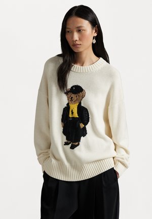 Maglione lavorato a maglia color crema con un orso ricamato che indossa un cappello nero, una camicia gialla e un cappotto nero. Polsini e orlo a coste.