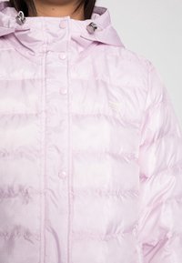 Ljusrosa puffer-jacka med huva, som har tryckknappar, kviltad textur och en liten logotyp på vänster sida.