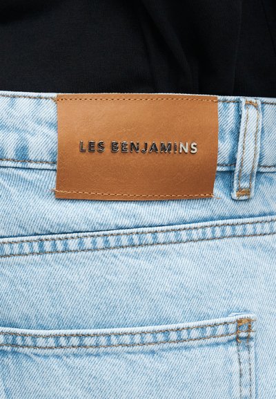 Jasnoniebieskie dżinsy z denimu z brązową skórzaną metką naszytą nad tylną kieszenią, tłoczoną napisem "LES BENJAMINS" w metalicznych literach.