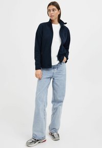 Giacca in pile navy con colletto alto, chiusura a zip e tasche laterali, abbinata a una t-shirt bianca e jeans a gamba larga di un blu chiaro.