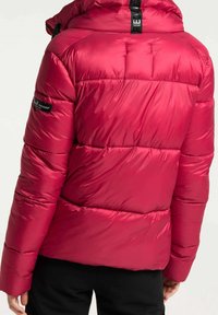 Rosa puffjacka med quiltad design, som har hög krage, elastiska ärmslut och en logotyp-patch på ärmen. Glansig, slät yta.