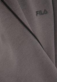 Tejido de mezcla de algodón gris con una textura suave. Presenta un logo de Fila en negro y una sutil costura de acento en negro. Diseño simple y moderno.