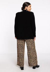 Vrouw die met haar rug naar de camera staat, draagt een zwarte langemouwen top, een brede broek met luipaardprint, zwarte hoge hakken en zwarte hangende oorbellen, terwijl ze naar de zijkant glimlacht.