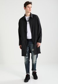 Sort letvægtsjakke med lige snit, der har en skjult stolpe og sidelommer, båret over en hvid T-shirt og slidte blå jeans.