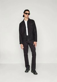 Tânăr purtând o șapcă neagră cu spatele, ochelari de soare negri, jachetă neagră, pantaloni negri, tricou alb și pantofi negri, stând pe un fundal deschis.