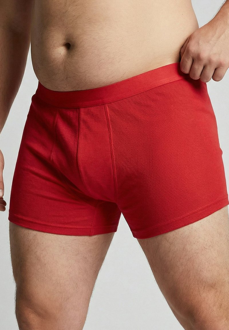 Torso masculin et cuisses supérieures portant un boxer rouge côtelé, la main droite ajustant la ceinture, sur fond uni.