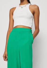 Bílý pruhovaný crop top s vysokým výstřihem, zkombinovaný s volnými zelenými kalhotami s pasem a bočními kapsami. Včetně dekoračního náhrdelníku.