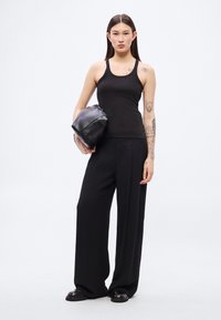 Μαύρο ριμπ τοπ με λεπτές τιράντες, συνδυασμένο με μαύρο παντελόνι φαρδιάς γραμμής. Κρατάει μια μαύρη τσάντα pouch. Απλός, μινιμαλιστικός σχεδιασμός και χρώματα.