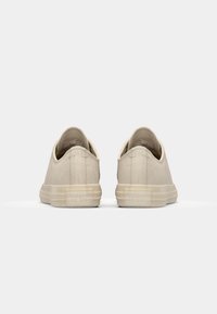 Zapatillas de lona beige con diseño de corte bajo, suelas de goma planas y un acabado texturizado. Presenta un logo en relieve en el talón.