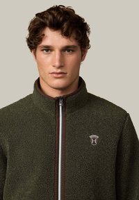 Tummanvihreä fleece-takki, jossa on korkea kaulus, ruskea vetoketju ja valkoinen raidallinen yksityiskohta. Edessä rintalogo kilpimallisena.